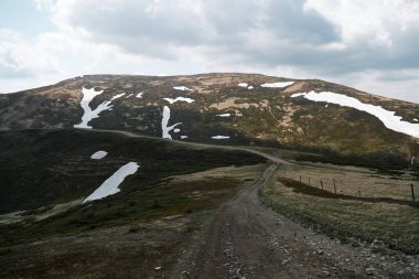 Kar ve çimlerle kaplı arazide dolambaçlı toprak yolu olan sakin dağ manzarası, yürüyüş ve doğa fotoğrafçılığı için ideal..
