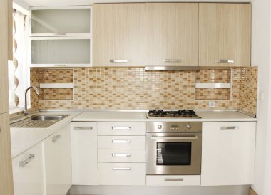 Mutfağın içi bej mobilyalarla, dolaplarla, raflarla ve yemek pişirmek için aletlerle dolu.