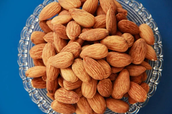 dried grains of natural brown almonds in a transparent decorative plate on a blue background