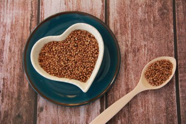 Kalp şeklinde dekoratif bir tabakta ve tahta bir kaşıkta küçük kahverengi karabuğday taneleri.