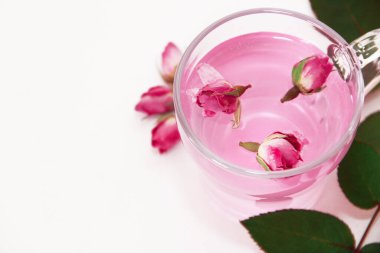 Şeffaf cam bardakta pembe çay ve beyaz arka planda pembe gül tomurcukları.