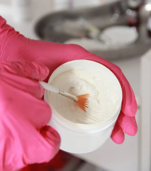 The hands of a cosmetologist doctor in pink gloves hold a brush and stir a white cream
