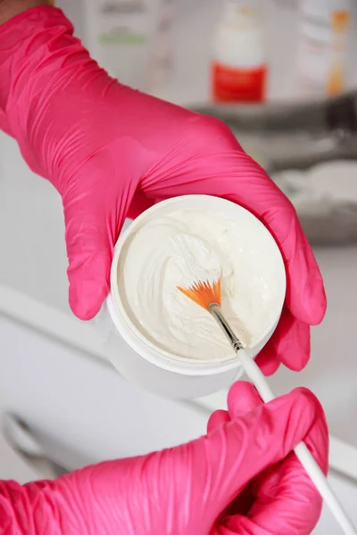 The hands of a cosmetologist doctor in pink gloves hold a brush and stir a white cream