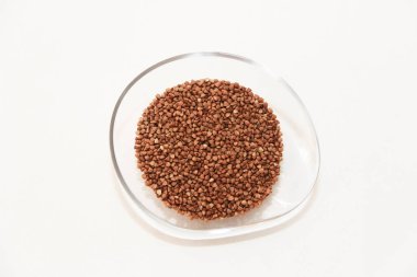 Small grains of natural brown buckwheat in a transparent plate on a light background.