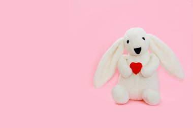 children's toy white plush bunny with long ears holds a red heart in its paws on a pink background