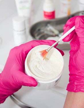 The hands of a cosmetologist doctor in pink gloves hold a brush and a white cream in a jar