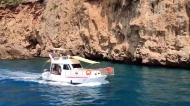 Mavi Akdeniz 'de kahverengi kayalardan oluşan bir arka planda beyaz bir motorlu tekne yüzüyor.