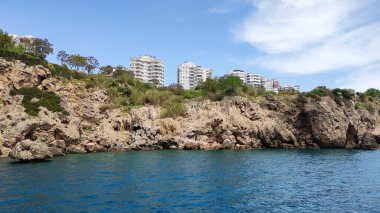 Mavi Akdeniz, mavi gökyüzü, beyaz bulutlar ve kayalık bir sahil.