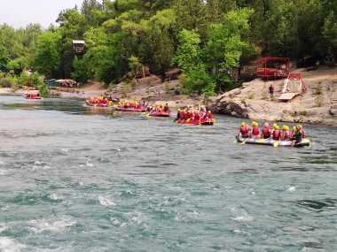Yelekli bir grup insan, resimli bir orman bölgesinde dağ nehri boyunca rafting tekneleriyle geziyorlar.