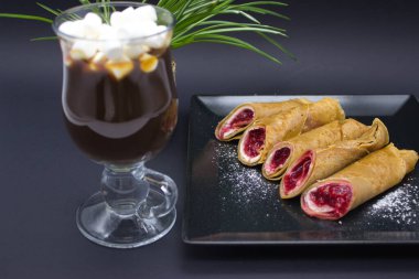 Ön planda, bu görüntü izleyiciyi baştan çıkarıcı bir bardak sıcak çikolata ile şımartıyor, üzeri şekerleme ve karamel serpiştiriyor. Arka planda, canlı bir böğürtlen kompostosu ile dolu altın krepler siyah bir tabakta tozla kaplıydı.