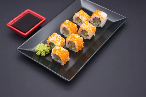 This image features a sleek composition of sushi, wasabi, and soy sauce on a dark, reflective surface. The sushi rolls, topped with bright orange masago, are presented in a line, leading the eye toward a red soy sauce dish in the background. A mound