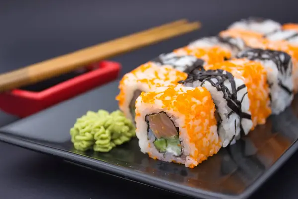 stock image This enticing image displays a row of California sushi rolls adorned with orange masago, arranged alongside a dollop of wasabi and a red dish of soy sauce on a sleek, black tray.