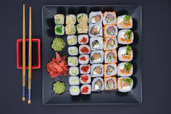 This vibrant image captures a variety of sushi rolls neatly displayed on a large black plate, accompanied by bamboo chopsticks, a red soy sauce dish, wasabi, and pickled ginger. The assortment includes rolls with avocado, fish, and vegetables, some w