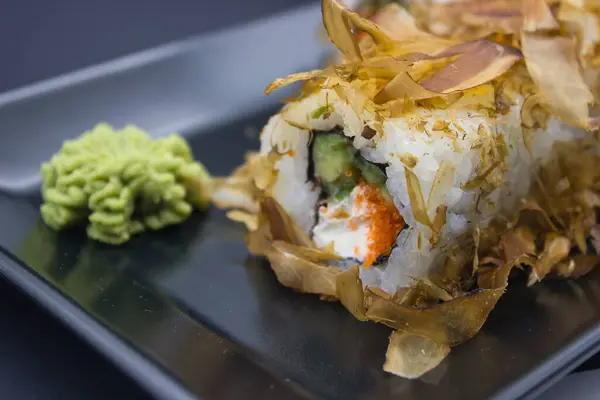 This close-up captures a sushi roll generously topped with bonito flakes, alongside a sculpted mound of wasabi, set on a reflective black tray. The textures of the flaked tuna, the creamy filling, and the crisp cucumber are vividly detailed, highligh