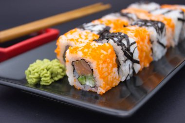 Bu cezbedici görüntü, turuncu masago ile süslenmiş bir sıra Kaliforniya suşi rulolarını, bir tutam wasabi ve parlak, siyah tepside kırmızı bir soya sosu ile birlikte gösteriyor..
