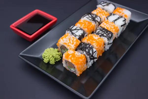 stock image This enticing image displays a row of California sushi rolls adorned with orange masago, arranged alongside a dollop of wasabi and a red dish of soy sauce on a sleek, black tray.