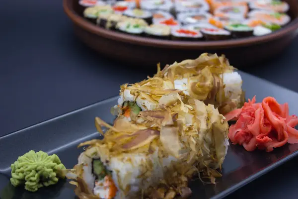 This close-up captures a sushi roll generously topped with bonito flakes, alongside a sculpted mound of wasabi, set on a reflective black tray. The textures of the flaked tuna, the creamy filling, and the crisp cucumber are vividly detailed, highligh