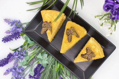 Taze çiçeklerle çevrili, çikolata kelebekleriyle süslenmiş üç ballı pasta dilimi..