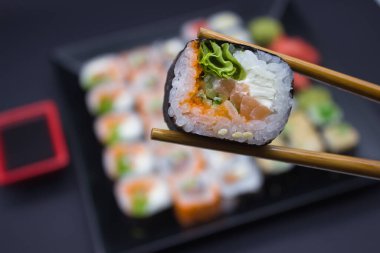 Yemek çubukları arasında tutulan büyüleyici bir suşi seçimi, siyah bir tabakta sergilenen çeşitli suşi türlerinden oluşan bir sıranın üzerinde duruyor. Sahne, kırmızı soya sosu tabağı ve yeşil wasabi tepeciği ile çerçevelenmiş, turşulu zencefil de bir miktar renk katıyor.. 