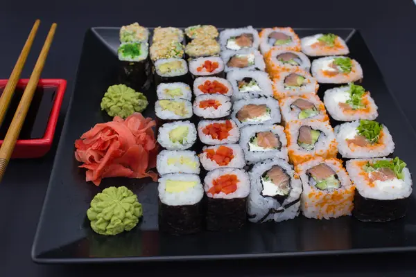 stock image This vibrant image captures a variety of sushi rolls neatly displayed on a large black plate, accompanied by bamboo chopsticks, a red soy sauce dish, wasabi, and pickled ginger. The assortment includes rolls with avocado, fish, and vegetables, some w