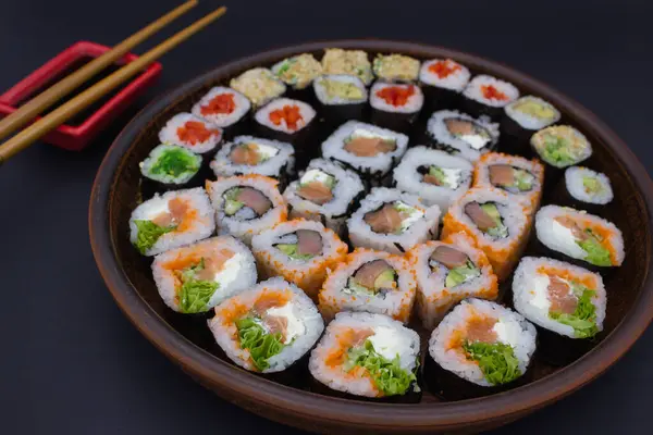 stock image This image features a large circular wooden platter filled with an array of sushi rolls, perfectly arranged in a circular pattern. The assortment includes a selection of rolls with fresh fish, avocado, and crisp vegetables. A small red dish filled wi