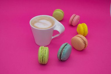 Renkli badem kurabiyeleriyle çevrili, parlak pembe arka planda bir fincan kahvenin yer aldığı rahat bir ortam..