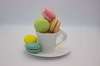 Beyaz bir fincanda zarif bir şekilde istiflenmiş renkli badem kurabiyesi çeşitleri. Düz beyaz arkaplandaki bu minimalist sunum bu Fransız şekerlerinin canlı renklerini ve hassas dokularını vurguluyor..