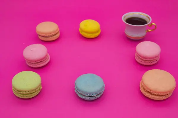 stock image An artistic arrangement of colorful macarons surrounding a delicate coffee cup on a vivid pink background, creating a delightful and eye-catching dessert presentation.