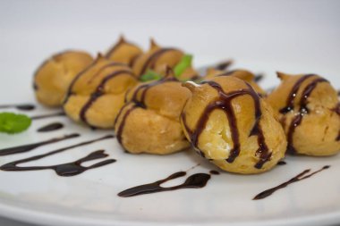 Yumuşak çikolata soslu krema dolu profiterollerin yakın plan görüntüsü. Altın hamur işleri taze nane yapraklarıyla vurgulanır. Bu da tatlıyı cezbedici bir ikram yapar..