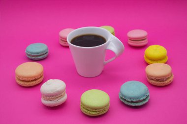 Canlı pembe bir zemin üzerinde bir fincan koyu kahveyi çevreleyen renkli badem kurabiyesi dairesi çarpıcı bir kontrast ve görsel olarak çekici bir sahne yaratıyor..