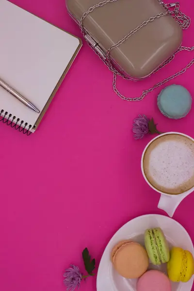 Stock image A stylish flat lay featuring colorful macarons on a plate, a frothy coffee, a chic purse with a chain, and a notebook with a pen on a bright pink background.