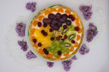 Canlı bir meyveli tartın bu yakın çekimi bir dizi sulu böğürtlen, üzüm, kivi ve kremalı dolgu üzerine dizilmiş bitki örtüsünü ön plana çıkarıyor. Jöle taze, ışıl ışıl bir doku ekler. Tatlının görsel çekiciliğini arttırır..