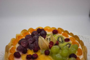 Canlı bir meyveli tartın bu yakın çekimi bir dizi sulu böğürtlen, üzüm, kivi ve kremalı dolgu üzerine dizilmiş bitki örtüsünü ön plana çıkarıyor. Jöle taze, ışıl ışıl bir doku ekler. Tatlının görsel çekiciliğini arttırır..