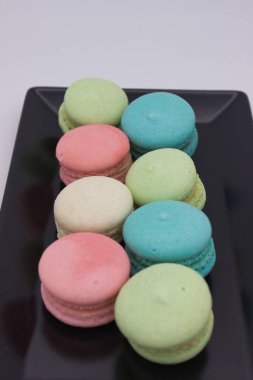 Siyah bir tabakta pastel renkli badem kurabiyelerinin yakın çekimi, canlı renklerini ve hassas dokularını gösteriyor. Görüntü bu klasik Fransız tatlılarının zarafetini ve çekiciliğini yakalıyor..