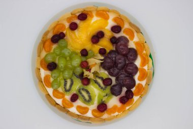 Canlı bir meyveli tartın bu yakın çekimi bir dizi sulu böğürtlen, üzüm, kivi ve kremalı dolgu üzerine dizilmiş bitki örtüsünü ön plana çıkarıyor. Jöle taze, ışıl ışıl bir doku ekler. Tatlının görsel çekiciliğini arttırır..