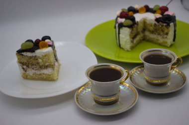 A cozy setting with a slice of fruit-topped layer cake on a white plate, a whole cake in the background on a green plate, and two cups of black coffee in elegant porcelain cups. clipart