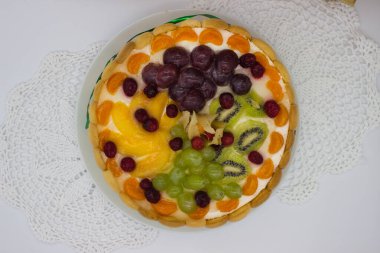 Canlı bir meyveli tartın bu yakın çekimi bir dizi sulu böğürtlen, üzüm, kivi ve kremalı dolgu üzerine dizilmiş bitki örtüsünü ön plana çıkarıyor. Jöle taze, ışıl ışıl bir doku ekler. Tatlının görsel çekiciliğini arttırır..