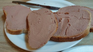 Birkaç dilim ekmeğin yakın plan çekimi, yumuşak karaciğer ezmesi ile güzelce yayılmış, arka planda bir bıçakla beyaz bir tabakta sunulmuştur.