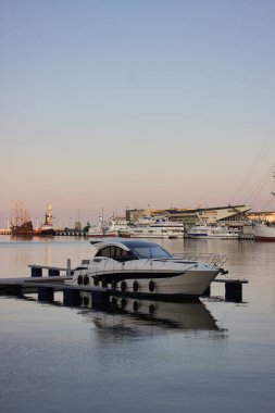Diğer gemiler ve günbatımı gökyüzü yansımalarıyla çevrili, deniz ya da seyahat temaları için ideal, sessiz marina suyunda yüzen modern beyaz motor yatı..