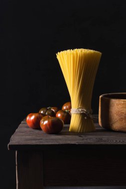 Some tomatoes and a bunch of spaghetti arranged vertically on an antique table. Dark background.