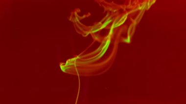 Sarı-yeşil mürekkep damlaları yükselir ve kırmızı arka ışıklandırmalı bir su kabında çözülür..