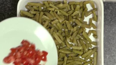 Beyaz bir tabaktan taze salata hazırlayan yeşil fasulyeli kare bir kaseye doğranmış kiraz domatesi eklenen ellerin yukarıdan aşağı bakışı..