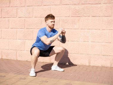 Atletik adam tuğla bir duvara karşı çömelmiş, dayanıklılık ve zindelik sergiliyor. Atletizm kıyafetleri giyiyor, açık havada formda ve dengede kalmaya odaklanıyor.