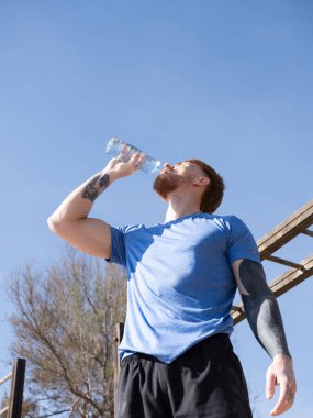 Kızıl saçlı sporcu güneşli bir günde açık havada spor salonunda antrenman yaptıktan sonra plastik şişeden su içiyor, spor yaparken dinleniyor ve su içiyordu.