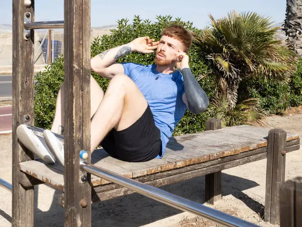 Odaklanmış sporcu, parktaki ahşap bir bankta oturarak vücut kaslarını güçlendirir. Bu da spor ve açık hava egzersizlerine olan bağlılığını gösterir.