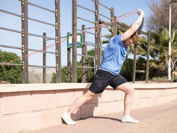 Genç sporcu, açık hava spor aletlerine bağlı direnç bandıyla egzersiz yapıyor güneşli bir günde esneme egzersizleri yapıyor, sağlıklı yaşam tarzını ve formülünü destekliyor.