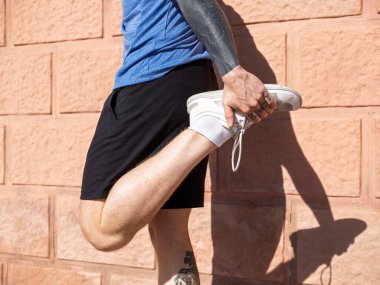 Atletik adam dışarıda tuğla duvara karşı bacak kaslarını esnetiyor. Spor kıyafetleri içinde güneşli bir günde antrenmana hazırlanıyor. Esneklik ve zindeliğe odaklanıyor.