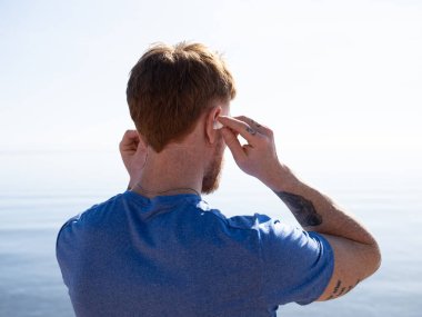 Okyanusun kenarında duran bir adam, dışarıda huzurlu bir anın tadını çıkarırken kulaklıklarını ayarlıyor. Kollarında dövmeler var, günlük mavi bir gömlek giyiyor, sakin sahil çevresini kucaklıyor.