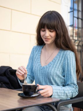 Serene genç kadın, açık bir kafede kahvesini karıştırıyor, canlı şehir atmosferinde huzurlu bir anın tadını çıkarıyor..