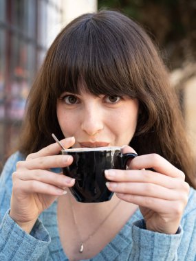 Genç bir kadın açık bir kafede sıcak bir içeceğin tadını çıkarıyor, espressonunun zengin aromasının tadını çıkarırken şehir sabahında dinleniyor.